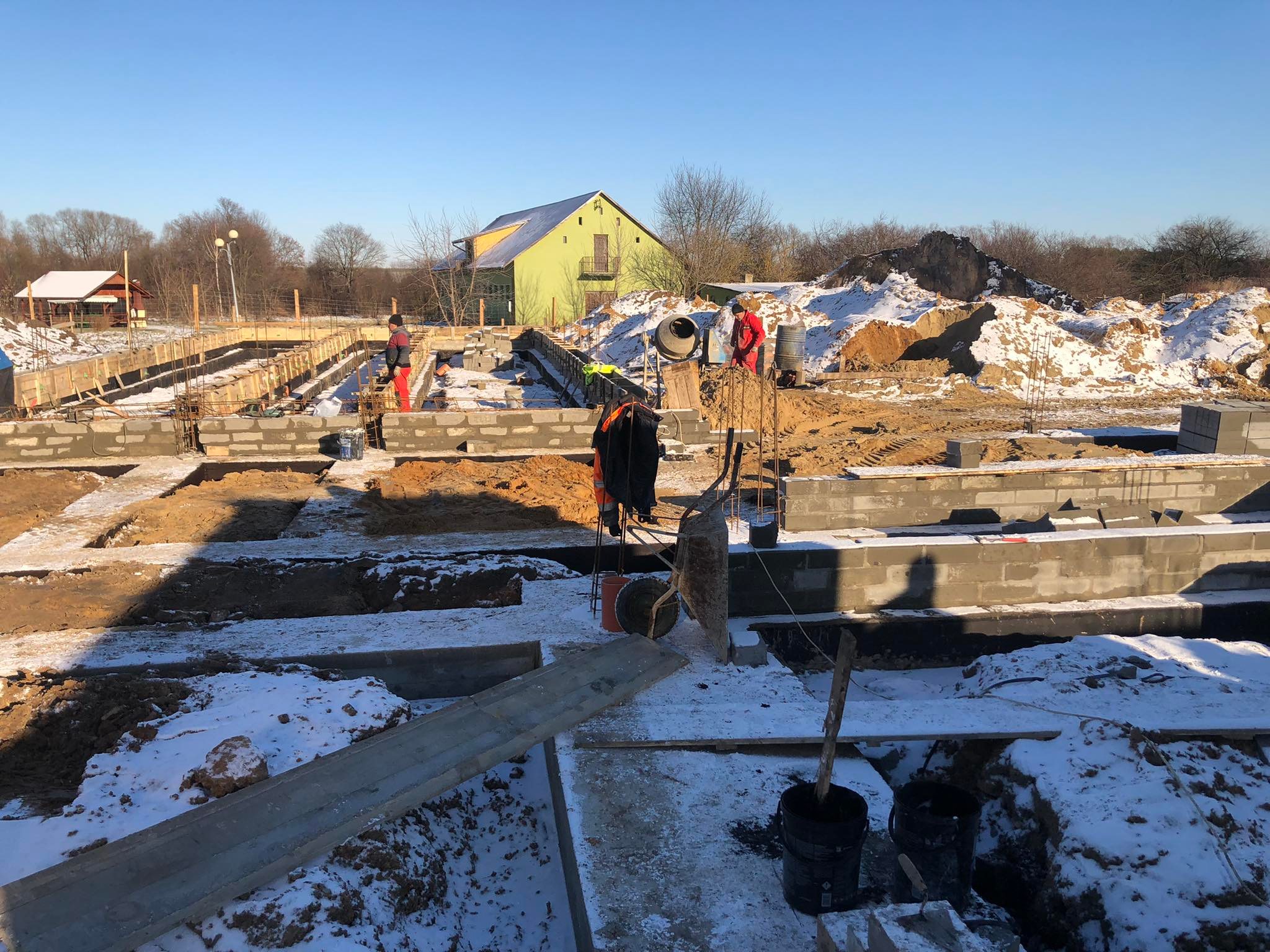 261673047_1200294147162761_1082299309863598588_n Plac budowy pokryty lekkim śniegiem. Robotnicy wykonują prace przy fundamentach.