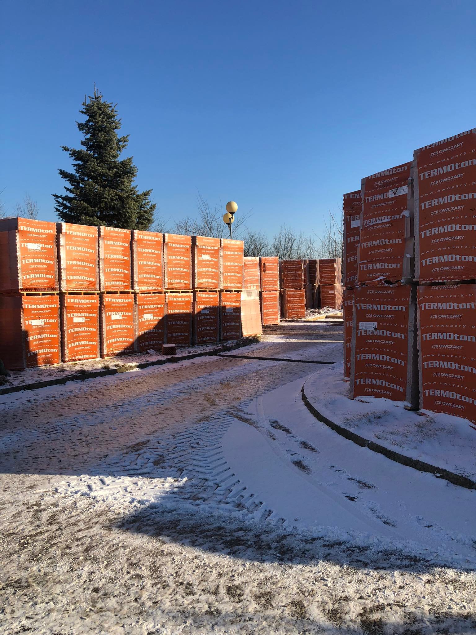 263192795_308732947808430_2964148200307239113_n Plac budowy pokryty lekkim śniegiem. Przy bramie wjazdowej stoją palety z materiałami budowlanymi.