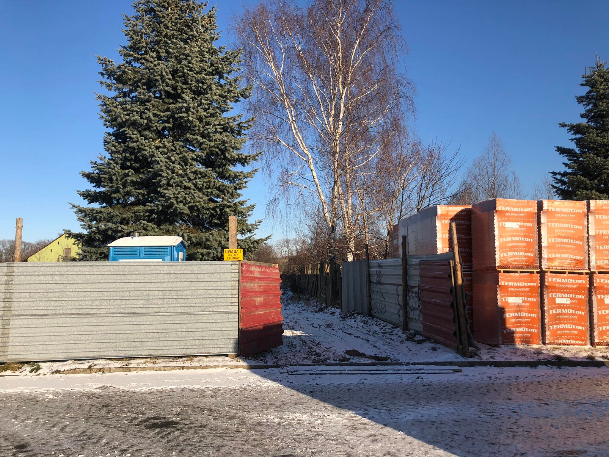 263733648_4783167728444730_2046483304629494210_n Plac budowy pokryty lekkim śniegiem. Przy bramie wjazdowej stoją palety z materiałami budowlanymi.